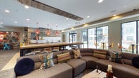 This single-story space presents an open-plan communal lounge designed with a blend of rustic and contemporary elements. A prominent bar-style counter runs along the back of the room, finished with a warm wood surface and backed by reclaimed wood paneling that adds a textured, industrial character to the space. Above the counter, a series of bold coral-red pendant lights hang at varying heights, providing both functional illumination and a striking visual focal point.

The ceiling incorporates recessed lighting alongside a structural wood beam detail that frames the kitchen and bar area, reinforcing the space's mixed-material aesthetic. The lounge area is anchored by a generously sized curved sectional sofa in a neutral brown upholstery, dressed with an array of colorful, patterned throw pillows that introduce lively energy to the seating zone.

Large floor-to-ceiling windows and glass doors line one wall, flooding the interior with natural light and offering direct visual connection to an outdoor courtyard area. The overall ambiance is welcoming and social, balancing comfort with purposeful design details that make the space feel both stylish and functional.