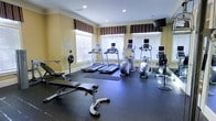 This fitness center offers a comprehensive workout environment designed to support a range of exercise routines. The space is finished with warm yellow walls, white crown molding, and recessed lighting, lending it a clean and polished aesthetic. Rubber flooring throughout the workout area provides both safety and durability underfoot.

The cardio section is well-stocked with multiple treadmills and an elliptical trainer, each equipped with individual monitors for entertainment or workout tracking. A rowing machine and an upright exercise bike round out the cardio offerings. For strength training, the space includes an adjustable weight bench with a barbell and additional free weight attachments, catering to a variety of lifting exercises.

Natural light fills the room through multiple windows dressed with striped fabric valances and horizontal blinds, creating a bright and energizing atmosphere. A wall-mounted flat-screen television provides additional entertainment options for those working out. Portable fans are positioned throughout the space to ensure comfort during intense sessions. The layout is thoughtfully organized, allowing ample room to move between equipment while maintaining an open, uncluttered feel. Overall, this fitness center provides a functional and inviting environment for daily exercise.
