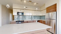 Located in Silver Spring, MD, this well-appointed 2-bedroom, 2-bathroom residence offers a thoughtfully designed single-level layout with modern finishes and an abundance of natural light. The open-concept kitchen is a standout feature, showcasing a large quartz-topped island with wood-grain cabinetry, a striking blue mosaic tile backsplash, stainless steel appliances, and pendant lighting with track fixtures overhead. The kitchen flows seamlessly into the living area, which is lined with floor-to-ceiling windows and a sliding glass door leading to a private balcony with views of the surrounding Silver Spring skyline and greenery. Light hardwood-style flooring runs throughout the main living areas, enhancing the bright, airy atmosphere. The spacious primary bedroom features plush carpeting, large windows framing outdoor views, and access to generous closet space with built-in wire shelving. The second bedroom is equally well-lit with wide windows and soft carpeting. Both bathrooms are finished with clean, contemporary tile work — one featuring a soaking tub and shower combination with a mosaic accent band and dark-toned vanity cabinetry. A central hallway connects all areas efficiently, and three closets provide ample storage throughout the residence.