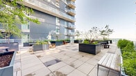 Located at 50 Place de l'Éclipse in Brossard, Québec, this beautifully designed rooftop terrace offers a generous open-air retreat for residents and guests. The terrace is finished with large-format stone pavers laid in a clean grid pattern, lending a refined, contemporary aesthetic to the space. Generous in scale, the terrace is thoughtfully organized to accommodate both social gatherings and quiet relaxation.

Large, deep navy blue planters are distributed throughout the space, filled with a variety of ornamental grasses, shrubs, flowering plants, and young trees that bring life and greenery to the elevated setting. Adirondack chairs in warm wood tones are positioned at various points around the terrace, inviting leisurely seating with open-sky views. Picnic-style tables with benches provide dedicated areas for group dining or casual conversation.

The terrace is bordered by glass railings that maintain unobstructed sightlines while ensuring safety, allowing visitors to take in the wide panoramic views of the Brossard skyline and surrounding urban landscape. The bold blue façade of the adjacent building frames parts of the terrace, reinforcing the modern architectural character of the development. This well-appointed outdoor space serves as a vibrant communal amenity, combining thoughtful landscaping with functional, comfortable furnishings.