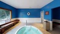 This spa room is defined by its striking bold blue upper walls and white-tiled lower wainscoting, creating a clean, aquatic aesthetic throughout the space. The centerpiece is a generously sized circular whirlpool tub set into the concrete floor, accessible via a handrail and offering ample room for relaxation. Recessed ceiling lighting illuminates the area, complemented by a large window along one wall that invites natural light and provides views of the surrounding outdoor greenery. A substantial dark-stained wooden bench runs along the window wall, offering a practical resting area, with additional wooden seating tucked into a corner alcove. The room includes standard safety signage and a life ring mounted on the wall, reinforcing the aquatic environment. A waste receptacle and posted rules are conveniently positioned near the entry. Two doorways provide access to adjoining areas, ensuring smooth circulation. The concrete flooring and tiled lower walls are practical and easy to maintain, while the overall design balances functionality with a resort-style ambiance suited to communal relaxation.