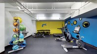 This single-level fitness facility offers a thoughtfully organized workout environment designed to accommodate a variety of exercise routines. The space features a bold, energetic aesthetic defined by a bright yellow accent wall and a vivid blue wall adorned with decorative circular motifs and a dolphin graphic, creating a lively and motivating atmosphere.

The room is outfitted with a selection of cardio equipment, including a treadmill and an elliptical machine, positioned along one side for easy access. A two-tier dumbbell rack along the yellow wall provides a range of free weight options, accompanied by a flat workout bench for strength training exercises. Along the opposite wall, an assortment of stability and exercise balls in various sizes are neatly stored on a rack, alongside rolled yoga mats resting on the floor nearby.

The ceiling features exposed white-painted pipes and overhead lighting, contributing to an industrial yet clean aesthetic. The flooring consists of dark rubber matting, providing durability and safety underfoot. A water fountain is mounted on the wall, ensuring easy hydration access. The overall layout maximizes the available floor space efficiently.