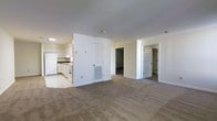 This single-floor space features an open-concept layout that seamlessly connects the kitchen and living areas. The kitchen is finished with white cabinetry, white appliances including a refrigerator and range, and light tile flooring, offering a clean and functional workspace. A wall with a built-in HVAC vent separates the kitchen from the broader living area, which is covered in neutral-toned wall-to-wall carpeting that extends throughout the main space and into the adjoining rooms. The walls are painted in a crisp white throughout, contributing to a bright and airy atmosphere. Two doorways visible from the main living area provide access to additional rooms, suggesting a multi-room floor plan suited for comfortable day-to-day living. The layout is straightforward and practical, with a clear delineation between the hard-surface kitchen zone and the softer carpeted living spaces. Overall, the unit presents a functional and well-organized interior with a neutral palette that offers a versatile canvas for personalization.