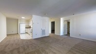 This single-floor space features an open-concept layout that seamlessly connects the kitchen and living areas. The kitchen is finished with white cabinetry, white appliances including a refrigerator and range, and light tile flooring, offering a clean and functional workspace. A wall with a built-in HVAC vent separates the kitchen from the broader living area, which is covered in neutral-toned wall-to-wall carpeting that extends throughout the main space and into the adjoining rooms. The walls are painted in a crisp white throughout, contributing to a bright and airy atmosphere. Two doorways visible from the main living area provide access to additional rooms, suggesting a multi-room floor plan suited for comfortable day-to-day living. The layout is straightforward and practical, with a clear delineation between the hard-surface kitchen zone and the softer carpeted living spaces. Overall, the unit presents a functional and well-organized interior with a neutral palette that offers a versatile canvas for personalization.