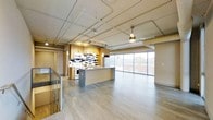 This two-floor urban loft combines industrial design with modern finishes to create a striking and functional living space. The main living area showcases an open-concept layout with light hardwood flooring that extends throughout, drawing the eye toward expansive floor-to-ceiling windows that flood the space with natural light and frame panoramic views of the surrounding cityscape.

The kitchen is a standout feature, outfitted with warm wood cabinetry, a mosaic tile backsplash in a bold geometric pattern, granite-topped counters, stainless steel appliances, and a central island that serves as a natural gathering point. Exposed ductwork, conduit, and track lighting run across the ceiling, reinforcing the loft's industrial character while maintaining a clean, contemporary feel. A ceiling fan with a brass finish adds a functional accent to the open living area.

A staircase connects the two floors, offering access to additional living space above. The upper level provides access to an outdoor rooftop terrace enclosed by a steel mesh railing, where sweeping views of the urban skyline — including a domed civic building in the distance — create a compelling backdrop. The overall ambiance balances raw industrial elements with refined modern touches, making for a dynamic and distinctly urban environment.