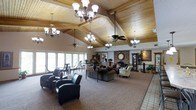 This single-story space is defined by its striking vaulted ceiling clad in warm, natural wood paneling supported by exposed timber beams, creating an inviting and airy atmosphere throughout. Multiple elegant chandeliers hang from the ceiling alongside ceiling fans, providing both ambient and functional lighting across the generous open floor plan.

The interior is organized into distinct seating areas, including a casual lounge zone with leather armchairs and upholstered sofas arranged around a central coffee table, as well as a more intimate dining or activity area near large windows along one wall. These expansive windows invite abundant natural light into the space and offer views of the surrounding exterior landscape.

A prominent brick fireplace serves as a focal architectural anchor at one end of the room, accompanied by a decorative wall clock and framed artwork that add character and warmth. Decorative plants and floral arrangements are positioned throughout, enhancing the welcoming ambiance. The flooring transitions between patterned carpet in the main seating areas and tiled surfaces toward the perimeter, further defining functional zones within this well-appointed communal gathering space.