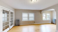This single-floor unit combines classic architectural character with practical living spaces throughout. Warm hardwood flooring flows continuously across the main living areas, lending a cohesive and inviting feel to the space. The main living room is highlighted by elegant multi-pane French doors that separate it from the adjoining room, while cast iron radiators serve as charming period details and provide reliable warmth. Multiple windows fitted with blinds allow natural light to filter in throughout the unit. An arched architectural detail adds a subtle decorative touch to the living area. The kitchen is well-appointed with rich dark wood cabinetry, granite countertops, a double-basin sink, and black appliances including a gas range and full-size refrigerator, all set over the same warm hardwood flooring. A secondary room features textured plaster walls and its own cast iron radiator beside a bright window. The bathroom showcases classic white subway tile with a decorative border accent, a built-in tub and shower combination, glass block windows that invite natural light while maintaining privacy, and a white vanity with sink. The entry area provides a clear transition point into the main living spaces, completing this well-structured and character-rich unit.