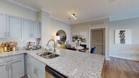 This single-floor residence in Kingsland, Georgia, presents a thoughtfully designed open-concept layout with refined finishes throughout. The kitchen serves as a central hub, featuring granite countertops, a large island with an undermount sink, soft gray shaker-style cabinetry, subway tile backsplash, and stainless steel appliances. Wood-style flooring flows through the main living areas, complementing the neutral color palette and crown molding details.

The living room offers a welcoming atmosphere with natural light streaming through large windows and glass doors that open to an outdoor view. The bedroom is generously proportioned, with plush carpeting, a bold accent wall, and a walk-in closet outfitted with wire shelving systems for ample storage organization.

A dedicated built-in workspace with a granite desktop surface and natural light provides a practical home office nook. The bathroom echoes the kitchen's design language with matching gray cabinetry and granite counters. A separate laundry area includes utility hookups, wire shelving, and wood-style flooring. The overall ambiance is modern yet comfortable, with cohesive finishes creating a polished, functional living environment.