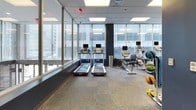 The Mayflower Fitness Center is a well-appointed facility designed to support a comprehensive workout experience. The entrance corridor features deep navy-blue walls, patterned carpet tile flooring, and glass partitions that provide a clear sightline into the main exercise area, creating an open and welcoming first impression.

The main workout floor is organized to accommodate both cardio and strength training. Along the perimeter, floor-to-ceiling windows flood the space with natural light and frame expansive city views, adding an energizing urban backdrop to every session. A row of treadmills, each equipped with individual monitors, lines the windowed wall, while elliptical trainers and a recumbent bike provide additional cardio options nearby.

The strength training area is equally well-equipped, featuring a dual cable machine, a dedicated leg extension/curl station, a full dumbbell rack ranging across multiple weights, and a flat bench for free weight exercises. Stability balls and medicine balls are also available, supporting functional and core training routines.

The space is finished with a drop ceiling fitted with recessed panel lighting, ensuring consistent illumination throughout. The neutral carpet tile flooring, combined with the clean architectural lines and dark accent walls, gives the fitness center a professional and polished atmosphere.
