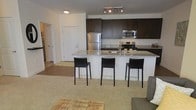 This single-floor space showcases an open-concept layout that seamlessly connects the kitchen and living areas. The kitchen is a functional centerpiece, featuring dark espresso cabinetry that contrasts elegantly with light granite countertops. Stainless steel appliances, including a refrigerator, electric range, and over-the-range microwave, provide a modern, polished aesthetic. A peninsula-style breakfast bar extends the counter space and serves as a natural dividing point between the kitchen and living area, accommodated by three sleek black bar stools. Track lighting above the kitchen ensures ample task illumination, complemented by recessed ceiling lights throughout. The adjoining living area is finished with neutral carpeting, contributing to a warm and inviting atmosphere. The open floor plan promotes a sense of spaciousness and ease of movement throughout the space. A hallway visible in the background suggests additional rooms beyond the main living area, offering functional separation of private and common spaces. Overall, the design balances practicality with contemporary style through its cohesive color palette and modern finishes.