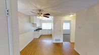 This single-floor residence offers a practical and well-connected layout designed for comfortable everyday living. The main living area is finished with warm hardwood-style flooring that extends throughout, creating a cohesive and inviting atmosphere. A ceiling fan with a light fixture is centrally mounted overhead, contributing to both airflow and ambient lighting across the space. The textured ceiling adds a classic residential character throughout.

The kitchen is open and accessible directly from the living area, featuring white cabinetry that lines the walls and provides ample storage. Dark countertops offer a subtle contrast to the bright cabinetry, and a window above the sink area allows natural light to filter in, brightening the workspace. The kitchen's clean, functional design makes it well-suited for daily use.

Adjacent to the main living space, a doorway leads to a carpeted bedroom, which benefits from its own window, bringing in additional natural light. A floor vent indicates a central HVAC system serving the space. The overall color palette is neutral throughout, with white walls and trim creating a bright, airy feel. The layout flows naturally between rooms, making this a straightforward and efficient single-floor living space.