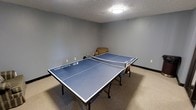 This single-floor space is designed with recreation and leisure in mind. The room is finished with neutral gray walls and wall-to-wall carpeting that adds a comfortable, casual feel underfoot. A textured ceiling with a centrally mounted flush light fixture provides steady, even illumination throughout the space. The focal point of the room is a full-size Eastpoint ping pong table in blue, complete with a net and paddles, making it a ready-to-use game area. A cushioned armchair is tucked into the corner, offering a comfortable spot for spectators or those taking a break between games. The room's simple, clean layout maximizes open floor space around the table, allowing for easy movement during play. Black baseboard trim outlines the perimeter of the room, adding a subtle finishing detail to the overall design. The space is practical and well-suited for recreational use, whether as part of a residential lower level, a community amenity room, or a dedicated game room within a larger facility.