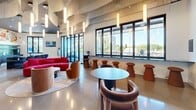 Located at 1600 Broadway in Sacramento's dynamic corridor, this single-floor amenity lounge is a thoughtfully designed communal space that blends contemporary architecture with eclectic artistic expression. The main lounge area is anchored by polished concrete floors, soaring ceilings with dramatic undulating ceiling installations, and floor-to-ceiling windows that flood the interior with natural light and offer sweeping views of the palm-tree-lined boulevard below. Pendant lighting cascades from above, complementing the open, airy atmosphere. A vibrant bar counter finished in teal, topped with a bold pop-art mural, serves as a focal point, flanked by sleek bar seating. Comfortable lounge furniture in rich reds and warm earth tones is arranged throughout, encouraging social interaction. A built-in fireplace with textured wall treatment adds warmth near the entrance area. The space connects to a generous outdoor terrace with modern metal railings, potted greenery, and seating that captures the energy of the Broadway streetscape. Two bathrooms continue the artistic theme — one featuring glossy blue-grey tile walls with an oval mirror and matte black sink, the other showcasing a colorful skateboard-inspired graphic tile accent wall, both finished with sleek fixtures and dark porcelain tile flooring.