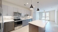 This one-bedroom, one-bathroom unit features a thoughtfully designed single-floor layout that maximizes functionality across a compact yet comfortable footprint. The kitchen serves as a standout centerpiece, showcasing two-tone cabinetry with light upper cabinets and dark lower cabinets, a classic subway tile backsplash, quartz countertops, and a full suite of stainless steel appliances including a refrigerator with ice maker, range, microwave, and dishwasher. A kitchen island with a wood-toned base and white quartz top provides additional prep space and is accented by stylish pendant lighting overhead. Light wood-tone flooring runs throughout the main living areas, lending a clean, contemporary feel. The living room benefits from natural light streaming through French doors that open to an outdoor area, complemented by a ceiling fan. The bedroom is carpeted for comfort, features a ceiling fan, and is filled with natural light from a window with blinds. It connects directly to a spacious walk-in closet outfitted with wire shelving and hanging rods for ample storage. The bathroom is finished with tile flooring, a dark wood vanity with a white countertop, a framed mirror, and a soaking tub with white subway tile surround.