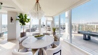 This expansive rooftop amenity level offers a remarkable blend of indoor sophistication and outdoor resort-style living, all elevated high above the city skyline with sweeping panoramic views of the surrounding urban landscape and distant mountain ranges.

The interior lounge area is defined by floor-to-ceiling glass walls that flood the space with abundant natural light while framing unobstructed city views. Light wood-toned flooring, a neutral palette, and carefully curated furnishings — including plush sectional sofas, accent chairs, and gold-toned decorative lighting — create a polished, contemporary atmosphere. A marble-accented feature wall adds a luxurious focal point, while a formal dining area with a round marble-top table, sculptural chandelier, and direct terrace access seamlessly connects indoor and outdoor living.

Outside, multiple outdoor pool decks are designed for resort-caliber relaxation. A sleek lap pool stretches along a sun-drenched terrace lined with chaise lounges, woven round daybeds with cushioned seating, pergola structures, and lush potted palms and agave plantings. Additional outdoor zones feature a spa, cabana-style seating areas, and manicured greenery. Clean-lined restroom facilities with ADA-compliant access serve the outdoor areas. Together, these amenities create an elevated, resort-inspired destination unlike any other.