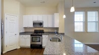 This single-story space is defined by thoughtful architectural details and a cohesive design aesthetic throughout. A hallway framed by a graceful arched opening sets the tone upon entry, drawing the eye toward the bright, open kitchen beyond. Dark hardwood flooring runs throughout, providing a rich contrast against the warm, neutral-toned walls and crisp white trim.

The kitchen is a standout feature, showcasing white cabinetry that extends to the upper walls, offering generous storage. Granite countertops with subtle speckled patterning provide durable, attractive work surfaces, continuing across a peninsula that includes an undermount stainless steel sink. Stainless steel appliances, including a range and an over-the-range microwave, complete the functional setup. Pendant lighting hangs above the peninsula, adding focused illumination and a decorative touch. Natural light enters through a window adjacent to the kitchen, brightening the space considerably.

Overall, the space presents a clean, move-in-ready environment with a balance of classic architectural elements and modern finishes, creating a functional and inviting atmosphere throughout.