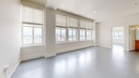 Located on the 12th floor at 11 E Forsyth Street in the heart of downtown Jacksonville, FL, this 3-bedroom unit offers an efficient and well-organized floor plan across a single level. The smooth, light gray epoxy flooring runs consistently throughout the space, lending a clean, modern aesthetic to every room.

The open-concept kitchen and living area forms the central hub of the unit. The kitchen features warm honey-toned wood cabinetry, a peninsula with a countertop overhang, and black appliances including a full-size refrigerator and dishwasher. The adjoining living room is bathed in natural light through a wide bank of windows fitted with horizontal blinds, offering sweeping elevated views of the Jacksonville skyline and surrounding cityscape.

The primary bedroom is generously proportioned, boasting corner windows on two walls that frame panoramic city views from multiple angles. A ceiling fan adds comfort, and a built-in closet provides practical storage. Additional bedrooms are bright and well-sized, with one featuring direct access to a dedicated closet with built-in shelving. The neutral, white-toned walls throughout create a clean and versatile backdrop, making the space highly adaptable to a variety of furnishing styles.