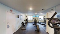 This single-level fitness space in Lancaster offers a dedicated workout environment designed for cardiovascular training. The room features a selection of cardio equipment, including a rowing machine, elliptical trainer, and treadmill, all positioned to maximize usability within the space. A full-length mirror spans one wall, visually expanding the room and allowing users to monitor their form during exercise. Two wall-mounted flat-screen televisions are positioned near the windows, providing entertainment during workouts. Natural light enters through multiple windows fitted with white blinds, brightening the space alongside recessed overhead lighting. The flooring consists of dark rubber gym matting, providing both durability and cushioning suited for high-impact activity. The walls are finished in a clean, light neutral tone, contributing to an open and energizing atmosphere. Ventilation is supported by a ceiling vent, ensuring adequate airflow throughout the room. The space is compact yet efficiently organized, making it a practical and functional facility for regular fitness routines.