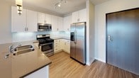 This single-floor unit offers a well-designed, open-concept layout that seamlessly connects the kitchen and living area. The kitchen is outfitted with white shaker-style cabinetry extending to the ceiling, dark quartz countertops, a subway tile backsplash, and a full suite of stainless steel appliances including a gas range, over-the-range microwave, dishwasher, and side-by-side refrigerator. A peninsula with a built-in sink and pendant lighting above defines the boundary between the kitchen and the spacious living area, which features wood-look plank flooring and large windows that invite abundant natural light and exterior views.

The bedroom is carpeted and includes a sizable window with outdoor views. An adjacent full bathroom offers a wide vanity with white cabinetry, dark hardware, tile flooring, and a tub-shower combination. A secondary bathroom with similar finishes is also present. A dedicated laundry area houses a stacked washer and dryer unit alongside wire shelving for additional storage, and connects to one of the bathrooms. Neutral-toned walls and consistent finishes throughout give the space a clean, cohesive, and modern aesthetic.