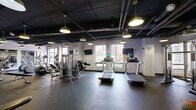 Located at 100 Wells St in downtown Hartford, Connecticut, this fitness center offers a comprehensive workout facility spread across a single, generously sized floor. The space is divided into two distinct areas: a main gym floor and an adjoining studio room, connected by glass-paneled partitions that create a sense of openness while defining each zone.

The main gym floor features durable rubber flooring and is outfitted with a wide array of cardio and strength equipment, including treadmills, ellipticals, cable machines, and free weights. Wall-mounted flat-screen televisions are positioned throughout for entertainment during workouts. Large windows along the perimeter flood the space with natural light and frame views of the Hartford skyline, adding an energizing urban backdrop.

The studio room, separated from the main floor by glass doors, features light hardwood flooring, wall-mounted ballet barres, and stability balls, making it well-suited for yoga, barre, stretching, or group fitness classes. Large windows continue into this room, maintaining the bright, airy atmosphere.

Throughout the facility, an industrial aesthetic is achieved through exposed black-painted ceilings with visible ductwork and piping, complemented by pendant dome light fixtures that provide warm, focused illumination. The overall design balances functionality with a modern, urban character.