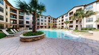 This expansive outdoor amenity area is the centerpiece of a multi-story residential complex, designed with a resort-inspired aesthetic that combines natural stone, warm wood, and manicured landscaping. The complex's U-shaped architectural layout wraps around the outdoor space, creating an enclosed, private courtyard atmosphere with balconies overlooking the grounds from multiple levels.

The swimming pool serves as the focal point, offering a generously sized, freeform design with a tanning ledge and in-water lounge chairs. The surrounding deck features stamped concrete pavers in warm earth tones, providing ample space for sunbathing and socializing. Tall palm trees and raised stone planter beds filled with colorful flowers add tropical character and visual interest throughout.

A dedicated lounge area with chaise chairs is arranged along the pool's edge, offering relaxed seating in an open-air setting. Adjacent to the pool, a well-maintained turf lawn provides additional outdoor recreation space, bordered by lush hedging and seasonal plantings.

A covered outdoor kitchen pavilion anchors one corner of the amenity area, featuring a built-in stainless steel gas grill set into a stone surround beneath a rustic timber-frame pergola roof. Outdoor dining furniture with a shaded umbrella completes the al fresco entertaining setup. The entire space benefits from abundant natural sunlight, creating a vibrant and inviting atmosphere.