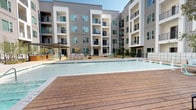 This outdoor pool area offers a well-appointed retreat designed for relaxation and recreation. The centerpiece is a generously sized swimming pool with crystal-clear water, framed by a warm wood-toned deck that provides ample space for lounging and socializing. A dedicated sun deck lines one side of the pool, furnished with rows of sleek white lounge chairs positioned to maximize sun exposure throughout the day.

Adding to the resort-like atmosphere, a series of private wood-framed cabanas line the poolside, each offering shaded seating and an elevated sense of comfort and privacy. A covered pergola area on one end of the pool provides additional shade and casual seating options for gatherings.

The pool area is enclosed within a courtyard formed by multi-story residential buildings featuring modern architectural design with clean lines, large windows, and private balconies. Mature trees and landscaped greenery are interspersed throughout, softening the hardscape and providing a natural, serene ambiance.

Access to the pool is provided through a dedicated entry point featuring rustic dark wood-framed doors set within a stucco exterior, accented by a wall-mounted lantern-style light fixture. A posted pool rules sign marks the entrance. The surrounding patio and concrete deck offer generous space for movement and relaxation, making this pool area a standout communal amenity.