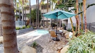 This outdoor courtyard is designed as a private resort-style retreat, centered around a spacious spa with tiled blue interiors and built-in hydrotherapy jets, accessible via a stainless steel handrail entry. The surrounding hardscape features stamped concrete patterned paving that extends into defined seating areas, furnished with wicker chairs and umbrella-shaded tables ideal for outdoor relaxation and socializing.

A dedicated outdoor kitchen station is built into one wall of the courtyard, equipped with two full-sized stainless steel BBQ grills mounted on a clean white countertop, making the space well-suited for outdoor entertaining. The area is enclosed by ornate wrought iron fencing with decorative finial detailing, providing both security and a refined aesthetic.

Tall, mature palm trees rise throughout the courtyard, lending a tropical ambiance and natural shade. The landscaping incorporates river rock ground cover, large decorative boulders, low ornamental grasses, tropical plants, and leafy shrubs, creating a layered and lush environment. Solar-style lanterns placed throughout add subtle accent lighting. The multi-story building surrounds the courtyard on multiple sides, creating a sheltered and private atmosphere that feels secluded despite its communal nature.