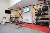 Located at 1830 Jerry Way in Norcross, Georgia, this fitness center is a well-appointed, single-floor facility designed to support a comprehensive workout experience. The space is organized into distinct functional zones, each tailored to a specific type of exercise.

The cardio area features a row of True-brand treadmills and additional cardio machines, including a stationary bike and elliptical. Multiple wall-mounted flat-screen TVs are positioned throughout for entertainment during workouts. Natural light flows in through a series of framed windows, complemented by bright linear overhead lighting that keeps the space well-lit and energizing.

A dedicated strength training zone is finished with red rubber flooring for impact absorption and includes a full dumbbell rack with medicine balls, a flat workout bench, and a striking whitewashed brick accent wall adorned with bold athletic mural graphics, adding a motivational, industrial character to the space.

The facility also includes a selection of cable and resistance machines for targeted strength training, positioned across neutral-toned carpet flooring. Large wall mirrors in the main area enhance the sense of openness and allow users to monitor their form. Ceiling fans provide additional air circulation, and the vaulted ceiling in portions of the space contributes to an airy, open feel. The overall design is clean, functional, and purpose-built for fitness.