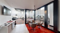 This fitness center in Denver, CO (80249) is a single-floor facility designed with both functionality and aesthetics in mind. The space is defined by dramatically high ceilings with exposed ductwork and industrial-style black ceiling panels, creating an open, energetic atmosphere. Rows of clerestory windows along the upper walls flood the interior with natural light while maintaining privacy.

The main workout floor is expansive, accommodating a comprehensive array of equipment across clearly defined zones. The cardio section features multiple TRUE-brand treadmills, stair climbers, and stationary bikes arranged in orderly rows. The strength training area includes a multi-station cable machine, Smith machine, free-weight benches, and a full dumbbell rack. A dedicated functional training zone offers TRX suspension systems, kettlebells, and medicine balls.

An artful geometric mural of a figure in motion decorates one wall, complementing the bold orange, gray, and white patterned carpet that runs throughout the space. A glass-and-cable partition wall separates a lounge area furnished with modern swivel chairs, offering a comfortable transition space. Wall-mounted televisions are positioned throughout for convenient viewing during workouts, enhancing the overall user experience.