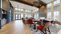 This beautifully designed single-floor community clubhouse delivers an upscale, resort-style atmosphere through thoughtful architecture and curated interior design. The centerpiece is a grand main lounge distinguished by soaring double-height ceilings and an expansive wall of floor-to-ceiling windows that flood the space with natural light and offer views of the surrounding outdoor amenity areas. A dramatic floor-to-ceiling stone-clad fireplace anchors the room, complemented by a striking sculptural gold chandelier and recessed ceiling details that add visual depth. Comfortable seating arrangements featuring neutral sofas, bold red accent chairs, and patterned area rugs create an inviting yet refined atmosphere. Wide-plank wood-look flooring flows throughout, unifying the various zones seamlessly. A secondary reception and lounge area showcases floor-to-ceiling open shelving with industrial black metal framing, curated sculptural décor, and warm leather and velvet furnishings. The adjacent game and entertainment room offers a stylish contrast, with a full-size pool table, bar-height communal seating, a large wall-mounted display screen, and a geometric black ceiling installation adding an industrial-modern edge. Together, these spaces form a dynamic, well-appointed clubhouse designed for both social gathering and relaxed leisure.