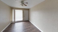This single-story unit is designed with a practical, functional layout that connects the living and kitchen areas in an open-concept arrangement. The living area features dark wood-style flooring, a ceiling fan with a light fixture, and a large sliding glass door fitted with vertical blinds, allowing natural light to fill the space and providing access to the exterior. Textured walls in a neutral beige tone are consistent throughout, contributing to a cohesive and understated aesthetic.

The kitchen is equipped with dark granite-style countertops that extend to form a peninsula, offering additional workspace and a natural divider between the kitchen and living areas. Rich, dark wood cabinetry lines the walls both above and below the counters, complemented by a stainless steel double-basin sink positioned beneath a window with blinds. A black refrigerator is also included.

The bedrooms are carpeted in a warm medium-tone brown, providing comfort underfoot. Each room features neutral walls, white baseboards, and windows with blinds that admit natural light. Ceiling fans are present in the bedrooms, enhancing air circulation. The overall ambiance of the unit is clean, neutral, and move-in ready.