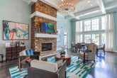 This single-story space in Buford, Georgia, presents a well-appointed interior defined by refined architectural details and a cohesive design aesthetic. Dark hardwood flooring extends throughout, providing a rich, grounding contrast to the soft blue-gray walls and bright white trim.

The living area serves as a central focal point, anchored by a striking floor-to-ceiling stone fireplace with a clean white mantel. Soaring ceilings enhance the sense of openness, while decorative crown molding and column accents add a layer of architectural sophistication. Bold abstract artwork adorns the walls, lending a contemporary, curated feel to the space.

The dining area is equally impressive, bathed in natural light streaming through an expansive bank of tall, multi-pane windows dressed with sheer patterned curtains. A French door provides direct access to the outdoor area, where views of the exterior grounds are visible. A substantial wood dining table with leather chairs anchors the room.

A dedicated workspace features a built-in countertop desk set against glossy white subway tile walls, complemented by warm pendant lighting and leather seating—a functional yet stylish area suited for productivity.