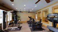 This fitness center offers a comprehensive workout environment designed to accommodate a range of exercise needs. The space is outfitted with an array of cardio equipment, including treadmills, elliptical trainers, and a recumbent bike, alongside a multi-station strength training machine and a dedicated free weights area with a weight bench. Protective rubber flooring mats are positioned beneath the equipment, providing both safety and durability. The ceiling features decorative wooden beam accents that lend a warm, rustic character to the space, complemented by natural wood trim along the walls and around the large framed mirrors on one side. The mirrors enhance the sense of openness while allowing users to monitor their form during workouts. Two ceiling fans keep air circulating throughout the room, and abundant recessed lighting ensures the space is well-lit at all times. Windows along one wall allow natural light to filter in, adding to the inviting atmosphere. The neutral-toned walls and carpeted areas outside the equipment zones create a comfortable and functional setting suitable for a full fitness routine.