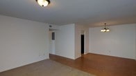This single-floor space offers a clean, functional layout designed for comfortable everyday living. The main living area features neutral white walls that create a bright, airy atmosphere, complemented by a flush-mount ceiling light fixture that provides warm illumination. Soft carpeting covers the living area, transitioning naturally into hardwood flooring that extends into the adjoining dining or secondary living space, adding a touch of warmth and visual contrast throughout. A partial wall divides the interior, creating a subtle separation between living zones while maintaining an open, connected feel. Doorways lead to additional rooms, hinting at further utility and storage beyond the main areas. The dining space is distinguished by a classic multi-bulb chandelier, lending a traditional character to the home. Visible HVAC vents in the ceiling reflect a practical, well-considered infrastructure. White baseboards run cleanly along the walls, reinforcing the space's simple, timeless aesthetic. Overall, this single-story layout balances openness with defined functional zones, making it a versatile and welcoming environment.