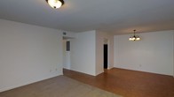 This single-floor space offers a clean, functional layout designed for comfortable everyday living. The main living area features neutral white walls that create a bright, airy atmosphere, complemented by a flush-mount ceiling light fixture that provides warm illumination. Soft carpeting covers the living area, transitioning naturally into hardwood flooring that extends into the adjoining dining or secondary living space, adding a touch of warmth and visual contrast throughout. A partial wall divides the interior, creating a subtle separation between living zones while maintaining an open, connected feel. Doorways lead to additional rooms, hinting at further utility and storage beyond the main areas. The dining space is distinguished by a classic multi-bulb chandelier, lending a traditional character to the home. Visible HVAC vents in the ceiling reflect a practical, well-considered infrastructure. White baseboards run cleanly along the walls, reinforcing the space's simple, timeless aesthetic. Overall, this single-story layout balances openness with defined functional zones, making it a versatile and welcoming environment.