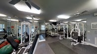 This fitness center offers a comprehensive workout environment designed to accommodate a range of exercise needs. The space is outfitted with multiple cable and weight machines positioned throughout the room, providing ample options for strength training. A dedicated free weight area features a full rack of dumbbells, while cardio equipment, including treadmills, is arranged nearby for convenience.

The flooring consists of durable rubber gym matting, providing both safety and comfort during workouts. Full-length mirrored walls line the perimeter, visually expanding the space and allowing users to monitor their form during exercise. Overhead, multiple ceiling fans work in tandem with bright flush-mounted lighting to keep the environment well-lit and comfortable.

Natural light filters into the space through windows visible along one wall, adding a welcoming brightness to the interior. A wall-mounted television is positioned for easy viewing from multiple workout stations. The overall layout is organized and functional, making efficient use of the available floor space while maintaining clear pathways between equipment.