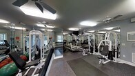 This fitness center offers a comprehensive workout environment designed to accommodate a range of exercise needs. The space is outfitted with multiple cable and weight machines positioned throughout the room, providing ample options for strength training. A dedicated free weight area features a full rack of dumbbells, while cardio equipment, including treadmills, is arranged nearby for convenience.

The flooring consists of durable rubber gym matting, providing both safety and comfort during workouts. Full-length mirrored walls line the perimeter, visually expanding the space and allowing users to monitor their form during exercise. Overhead, multiple ceiling fans work in tandem with bright flush-mounted lighting to keep the environment well-lit and comfortable.

Natural light filters into the space through windows visible along one wall, adding a welcoming brightness to the interior. A wall-mounted television is positioned for easy viewing from multiple workout stations. The overall layout is organized and functional, making efficient use of the available floor space while maintaining clear pathways between equipment.