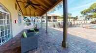 This single-story community clubhouse presents an inviting and well-appointed gathering space designed for comfort and leisure. The exterior is finished in warm yellow stucco, framed by a hip roof with dark shingles and accented by mature trees that provide natural shade and a pleasant, resort-like atmosphere.

A generously sized covered patio serves as the focal point of the space, featuring exposed wood-paneled ceilings supported by square columns and equipped with ceiling fans to ensure comfortable airflow. The patio floor is finished in richly textured brick pavers that extend throughout the outdoor areas, lending a cohesive and polished aesthetic. Wicker lounge chairs with green cushions and a low coffee table are arranged to create a relaxed seating area ideal for socializing or unwinding outdoors.

Elegant white-framed French doors with large glass panes and arched transom windows connect the covered patio to the interior of the building, allowing abundant natural light to flow through while offering views of the surrounding pool area and landscaping. White railing fencing borders the perimeter of the patio, providing a clean visual boundary between the clubhouse and the adjacent pool deck, where lounge chairs and umbrellas are visible. The overall ambiance is bright, airy, and tropical, making this a comfortable and functional communal destination.
