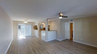 This single-floor residence offers a clean, open-concept layout that seamlessly connects the living area to the kitchen, creating a sense of spaciousness and ease of movement. Light wood-grain flooring runs throughout the main living area, lending a warm yet contemporary aesthetic.

The kitchen is partially open to the main living space, separated by a half-wall peninsula with a stone countertop that provides additional workspace and serves as a casual breakfast bar. Inside the kitchen, warm-toned backsplash tile adds texture and visual interest, while stainless steel appliances and white cabinetry with pendant lighting complete the functional yet stylish setup.

The living area benefits from generous natural light streaming through a large sliding glass door dressed with vertical blinds, which likely leads to an outdoor patio or balcony. A ceiling fan centered in the room provides both comfort and an additional design element.

Crisp white walls and clean white trim throughout contribute to a fresh and airy atmosphere. A doorway off the main living space leads to additional rooms, suggesting a well-organized floor plan. Recessed lighting and a HVAC vent integrated into the ceiling reflect thoughtful, modern utility. Overall, the space presents a welcoming, move-in-ready environment with a cohesive and practical design.