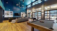 Located at 555 5th Ave. in Columbus, Ohio, this single-floor amenity space is a masterclass in contemporary community design. The centerpiece of the space is a dramatic double-height lounge area finished in a bold teal, with clerestory windows that flood the room with natural light. Warm hardwood flooring runs throughout, complemented by slatted wood accent columns and ceilings that add texture and visual interest.

The main lounge area features a regulation billiards table as a focal point, surrounded by ample open floor space ideal for social gatherings. Comfortable seating arrangements, including a large sectional sofa, accent chairs, and round side tables, create inviting conversation areas. Built-in white shelving and dark cabinetry line the teal accent wall, housing a wall-mounted flat-screen TV.

Adjacent to the main lounge is a separate relaxation area with a generously sized sectional sofa, a sputnik-style chandelier, and additional TV viewing. A sleek wet bar with a white quartz countertop, dark vertical-slat paneling, upholstered teal bar stools, and open wood shelving against a white tile backsplash serves as an elegant entertaining hub. A carpeted corridor lined with wood slat partitions connects the various zones, completing this well-appointed communal retreat.