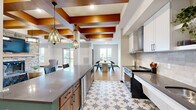 Located in Rosemount, Minnesota, this thoughtfully designed community space seamlessly blends a fully equipped kitchen with an expansive living area across a single open floor plan. The architecture immediately impresses with a signature coffered ceiling of rich warm wood beams set against crisp white panels, lending a refined yet welcoming character to the entire space.

The kitchen is a functional showpiece, featuring white upper cabinetry, a subway tile backsplash, stainless steel appliances, and a striking teal-accented island with a sleek gray quartz countertop and bar seating for several guests. Distinctive star-patterned floor tile anchors the kitchen zone, while pendant lights with amber glass add a warm, inviting glow overhead.

The living area flows naturally from the kitchen, centered around a full-height stacked-stone fireplace with a wood mantel and mounted television — a commanding focal point in the room. Light hardwood flooring extends throughout, complemented by a textured grasscloth feature wall that adds warmth and dimension. Multiple seating arrangements offer versatility, and large windows along the perimeter fill the space with natural light while providing views of the outdoor pergola area. The result is a cohesive, sophisticated environment ideal for gathering and entertaining.