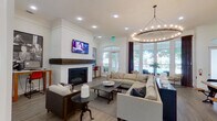 This single-story clubhouse-style space is thoughtfully designed to serve as both a social hub and a functional amenity center. The entry foyer sets an inviting tone with frosted glass doors, arched windows framed by dark curtain panels, and light hardwood-style flooring that extends throughout the entire space. A staffed reception desk with a bold pendant light featuring a yellow interior accent greets visitors upon arrival, with decorative columns adding architectural interest nearby.

The main lounge area is the centerpiece of the space, anchored by a white-painted fireplace with a thick, dark wood mantle and a wall-mounted television above it. A large sectional sofa and coffee table create a comfortable gathering area beneath a striking circular chandelier adorned with exposed Edison-style bulbs. Multiple arched windows and full-length glass doors flood the room with natural light and offer views of the surrounding greenery and outdoor pool area.

Adjacent to the lounge, a rustic communal dining table with industrial-style benches provides a dedicated space for group gatherings. A compact kitchenette nearby features dark wood cabinetry, white quartz countertops, open floating shelves, a stainless steel sink, and a beverage refrigerator. A workstation area with counter seating rounds out the functional offerings of this well-appointed space.