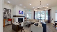 This single-story clubhouse-style space is thoughtfully designed to serve as both a social hub and a functional amenity center. The entry foyer sets an inviting tone with frosted glass doors, arched windows framed by dark curtain panels, and light hardwood-style flooring that extends throughout the entire space. A staffed reception desk with a bold pendant light featuring a yellow interior accent greets visitors upon arrival, with decorative columns adding architectural interest nearby.

The main lounge area is the centerpiece of the space, anchored by a white-painted fireplace with a thick, dark wood mantle and a wall-mounted television above it. A large sectional sofa and coffee table create a comfortable gathering area beneath a striking circular chandelier adorned with exposed Edison-style bulbs. Multiple arched windows and full-length glass doors flood the room with natural light and offer views of the surrounding greenery and outdoor pool area.

Adjacent to the lounge, a rustic communal dining table with industrial-style benches provides a dedicated space for group gatherings. A compact kitchenette nearby features dark wood cabinetry, white quartz countertops, open floating shelves, a stainless steel sink, and a beverage refrigerator. A workstation area with counter seating rounds out the functional offerings of this well-appointed space.