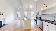 This single-floor unit offers an efficiently designed open-plan layout that combines the kitchen and living areas into one cohesive space. Soaring ceilings amplify the sense of roominess, while multiple large, black-framed windows flood the interior with natural light and provide views of the surrounding neighborhood. Light hardwood floors run continuously throughout, creating a warm, unified aesthetic.

The kitchen is thoughtfully appointed with white shaker cabinetry extending to the ceiling, dark stone countertops, a white subway tile backsplash, and a full suite of stainless steel appliances including a range, microwave, dishwasher, and a large bottom-freezer refrigerator. A central island with a matching dark countertop provides additional prep and seating space. Pendant lighting and a candelabra-style chandelier add character overhead.

The living area features a decorative arched fireplace with an ornate white surround and a black marble hearth, serving as a charming focal point. A ceiling fan completes the space.

The bathroom is well-appointed with herringbone-tiled shower walls, black penny-tile flooring, a teal vanity with a dark stone top, an arched mirror, and an in-unit stacked washer and dryer.