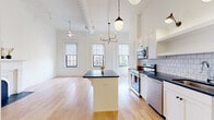 This single-floor unit offers an efficiently designed open-plan layout that combines the kitchen and living areas into one cohesive space. Soaring ceilings amplify the sense of roominess, while multiple large, black-framed windows flood the interior with natural light and provide views of the surrounding neighborhood. Light hardwood floors run continuously throughout, creating a warm, unified aesthetic.

The kitchen is thoughtfully appointed with white shaker cabinetry extending to the ceiling, dark stone countertops, a white subway tile backsplash, and a full suite of stainless steel appliances including a range, microwave, dishwasher, and a large bottom-freezer refrigerator. A central island with a matching dark countertop provides additional prep and seating space. Pendant lighting and a candelabra-style chandelier add character overhead.

The living area features a decorative arched fireplace with an ornate white surround and a black marble hearth, serving as a charming focal point. A ceiling fan completes the space.

The bathroom is well-appointed with herringbone-tiled shower walls, black penny-tile flooring, a teal vanity with a dark stone top, an arched mirror, and an in-unit stacked washer and dryer.