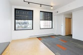 This single-floor unit presents a thoughtfully designed open-concept layout with a clean, contemporary aesthetic throughout. The kitchen is outfitted with light maple cabinetry, dark countertops accented by a mosaic tile backsplash, and stainless steel appliances including a refrigerator, gas range, microwave, and dishwasher. Cork-style flooring runs through the kitchen and entry areas, lending warmth to the space.

The main living area transitions to a patterned carpet tile floor in shades of grey and orange, adding a modern design element underfoot. Track lighting runs along the ceiling, providing flexible, directed illumination throughout. Two large, black-framed windows bring in generous natural light and offer urban cityscape views, enhancing the open feel of the living space.

A dedicated closet discreetly houses a stacked washer and dryer unit behind sliding maple-panel doors, adding practical convenience. The full bathroom features a combined tub and shower, a light wood vanity with dark countertop, and a distinctive black-and-white mosaic tile floor. The overall design is cohesive and modern, balancing functional storage with an airy, minimalist aesthetic well-suited to urban living.