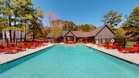 Located at 6350–6400 Oakley Rd in Union City, Georgia, The Oakley presents a well-appointed outdoor amenity space centered around a generously sized swimming pool with clear, aqua-blue water. The pool area is fully enclosed by a black iron fence, providing both security and an open, airy feel that connects seamlessly with the surrounding natural landscape of towering Georgia pines and mature trees.

The expansive concrete sundeck surrounds the pool on all sides, offering ample space for lounging and recreation. Bold red lounge chairs, chaise loungers, and umbrella-shaded seating arrangements are distributed throughout the deck, creating a vibrant and inviting atmosphere under the open sky. Multiple red patio umbrellas provide shaded gathering spots for outdoor dining and relaxation.

Adjacent to the pool, a modern clubhouse anchors the space architecturally. The building features a steeply pitched dark roof, board-and-batten siding, and large windows that allow natural light to flow through the interior. The combination of the contemporary clubhouse design and the lush, wooded backdrop gives the outdoor space a resort-like ambiance. Landscaped planting beds with shrubs and seasonal foliage line the perimeter, adding natural texture and color. The overall setting offers a comfortable, community-focused outdoor environment with a clear emphasis on leisure and outdoor living.
