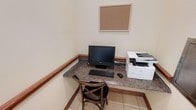 This single-floor business center is a focused, functional space designed to support productivity and convenience. The room features a built-in L-shaped granite countertop workstation with a rich, speckled stone surface that adds a polished, professional aesthetic. The workstation is outfitted with a desktop computer and keyboard, a mouse, and a multifunction printer, providing all the essentials for printing, scanning, and general computer use. A classic wooden cross-back chair is positioned at the desk, offering comfortable seating during work sessions. A cork bulletin board is mounted on the wall above the workstation, serving as a practical surface for pinning notes, schedules, or announcements. The walls are finished in a neutral, warm beige tone, complemented by warm wood baseboards that run along the perimeter of the room, lending a cohesive and welcoming feel to the space. The light-toned tile flooring is clean and easy to maintain, contributing to the overall bright and orderly atmosphere. The space is efficiently laid out to maximize utility within a compact footprint, making it well-suited as a shared business center in a hospitality, residential, or professional setting.