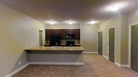 This single-floor space is designed with an open-concept layout that seamlessly connects the kitchen and living area, creating a functional and cohesive environment. The kitchen is equipped with dark espresso-toned cabinetry that provides ample storage, complemented by black appliances including a refrigerator, electric range, and overhead microwave. A peninsula-style breakfast bar with a granite-look countertop extends into the living space, integrating a built-in sink and offering additional workspace or casual seating. The flooring transitions from hard surfaces in the kitchen to neutral-toned carpet throughout the living area, adding warmth underfoot. Flush-mount ceiling light fixtures provide consistent overhead lighting across the open space. The walls are finished in a soft neutral tone, contributing to a clean and understated aesthetic. Two doorways visible at opposite ends of the space suggest access to additional rooms or exterior entry points, reinforcing the practical flow of the layout. White baseboards provide a crisp contrast against the carpeted floors and painted walls throughout.