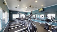 This fitness center is thoughtfully designed to support a well-rounded workout routine. The space is outfitted with a row of cardio machines, including treadmills and ellipticals, positioned to take advantage of natural light streaming in through a series of arched windows along the walls. The windows not only brighten the interior but also create an open, airy atmosphere that enhances the exercise experience.

A floor-to-ceiling mirror spans a full wall, allowing users to monitor their form while also visually expanding the space. Adjacent to the cardio area, a strength training station provides additional equipment for resistance-based workouts. The flooring is a durable, dark rubberized material well-suited for high-impact activity.

The ceiling features recessed lighting and dual ceiling fans, ensuring consistent air circulation and a comfortable environment throughout the room. A wall-mounted flat-screen television adds an entertainment element for those looking to stay engaged during longer workout sessions.

The walls are painted in a bold teal tone, lending the space an energetic and motivating ambiance. Crown molding along the ceiling adds a polished architectural detail, elevating the overall finish of the fitness center beyond purely functional design.